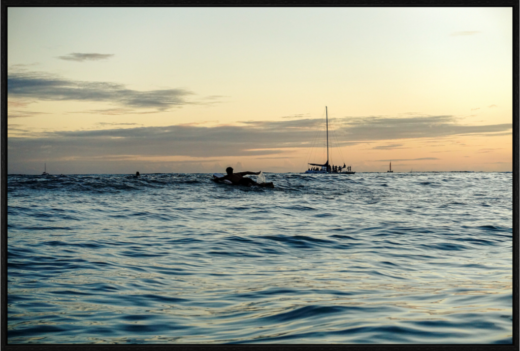 Surfer paddling out at sunset with catamarans in the distance