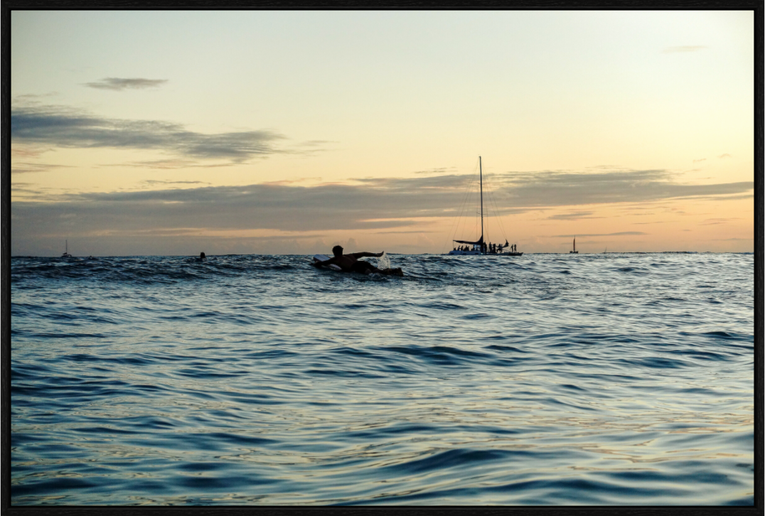 Surfer paddling out at sunset with catamarans in the distance